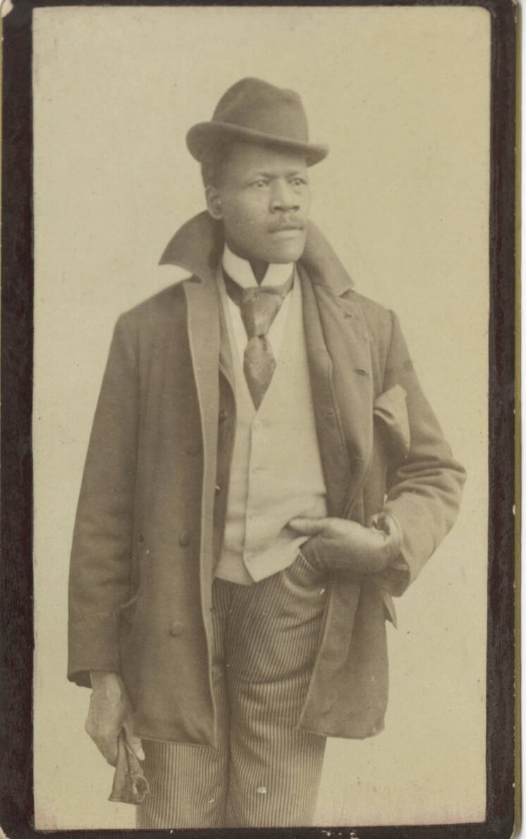 Photographic portrait of a man wearing a tie, hat, and jacket. His had his left hand partially in his pants pocket.